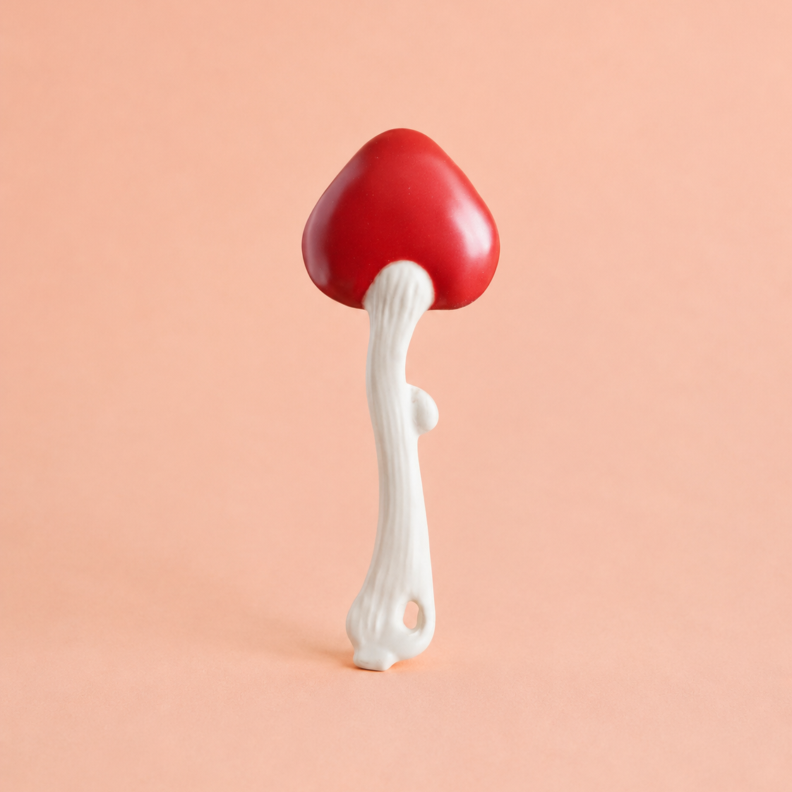 A mushroom-shaped spoon with a glossy red cap and a textured white stem, standing upright against a solid peach-pink background.