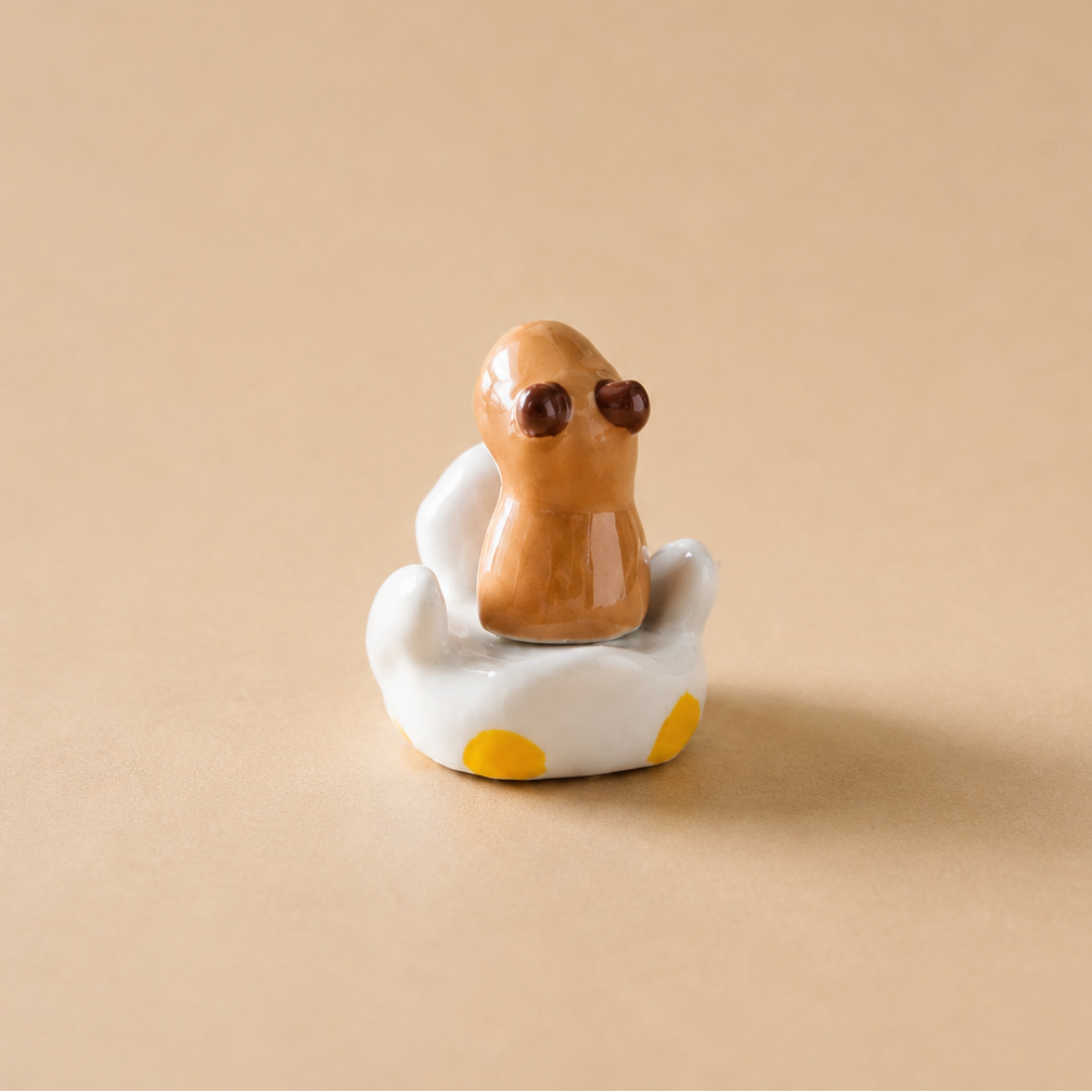 A small ceramic figurine of a brown capybara sitting in a white clam shell with yellow accents, set against a solid beige background.