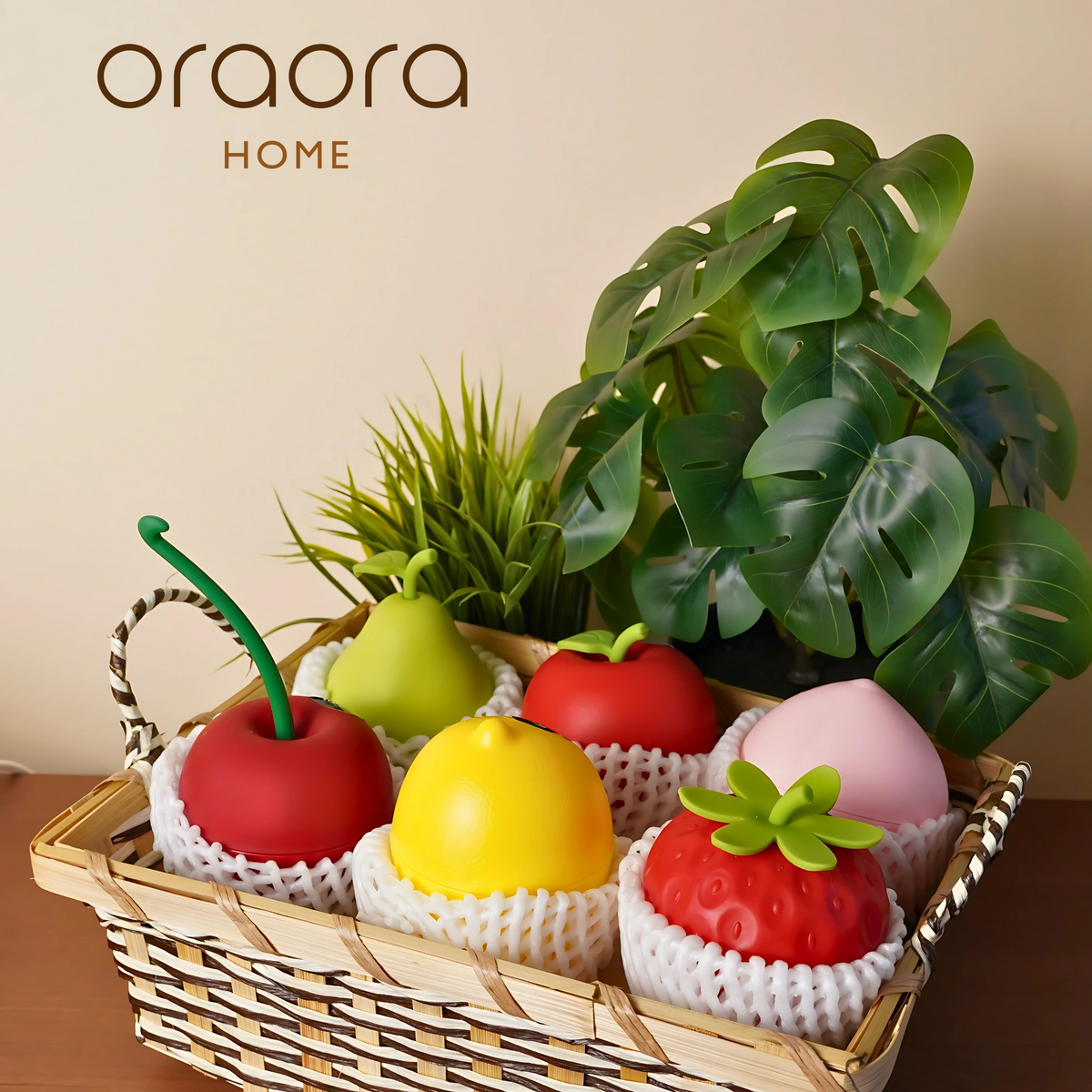A set of fruit-shaped aromatherapy air fresheners in a wicker basket, with a faux green plant to the left. The air fresheners are designed to look like apples, lemon, and other fruits.
