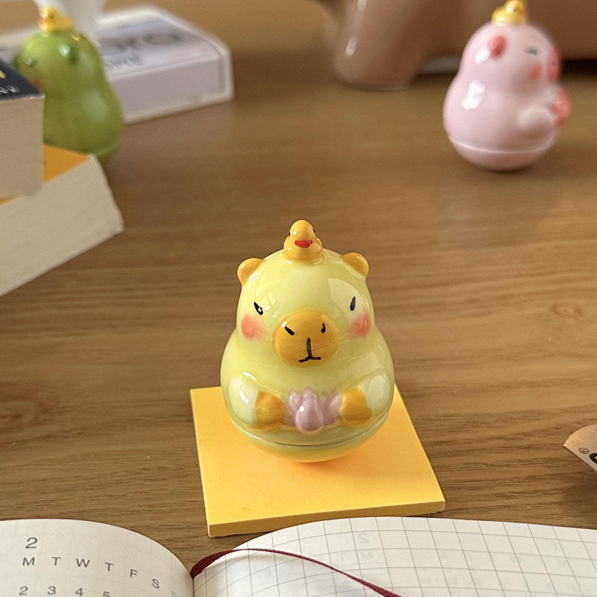 Small Capybara Desk Toy on a wooden surface next to an open notebook and calendar.