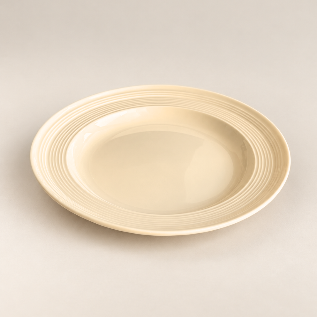 A round ceramic plate in a warm cream or ivory color, featuring a flared rim with concentric ridges, photographed on a neutral light gray background.