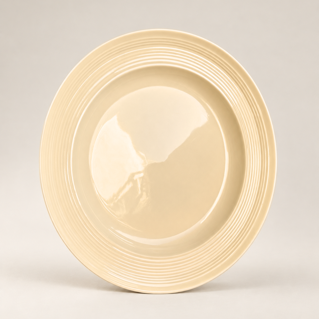 A round ceramic plate in a warm cream or light beige color, featuring a smooth glossy center and a flared rim with concentric ridges, photographed against a neutral light gray background.