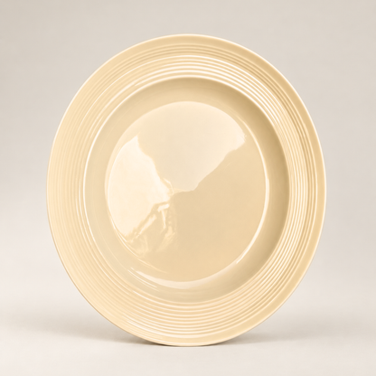 A round ceramic plate in a warm cream or light beige color, featuring a smooth glossy center and a flared rim with concentric ridges, photographed against a neutral light gray background.