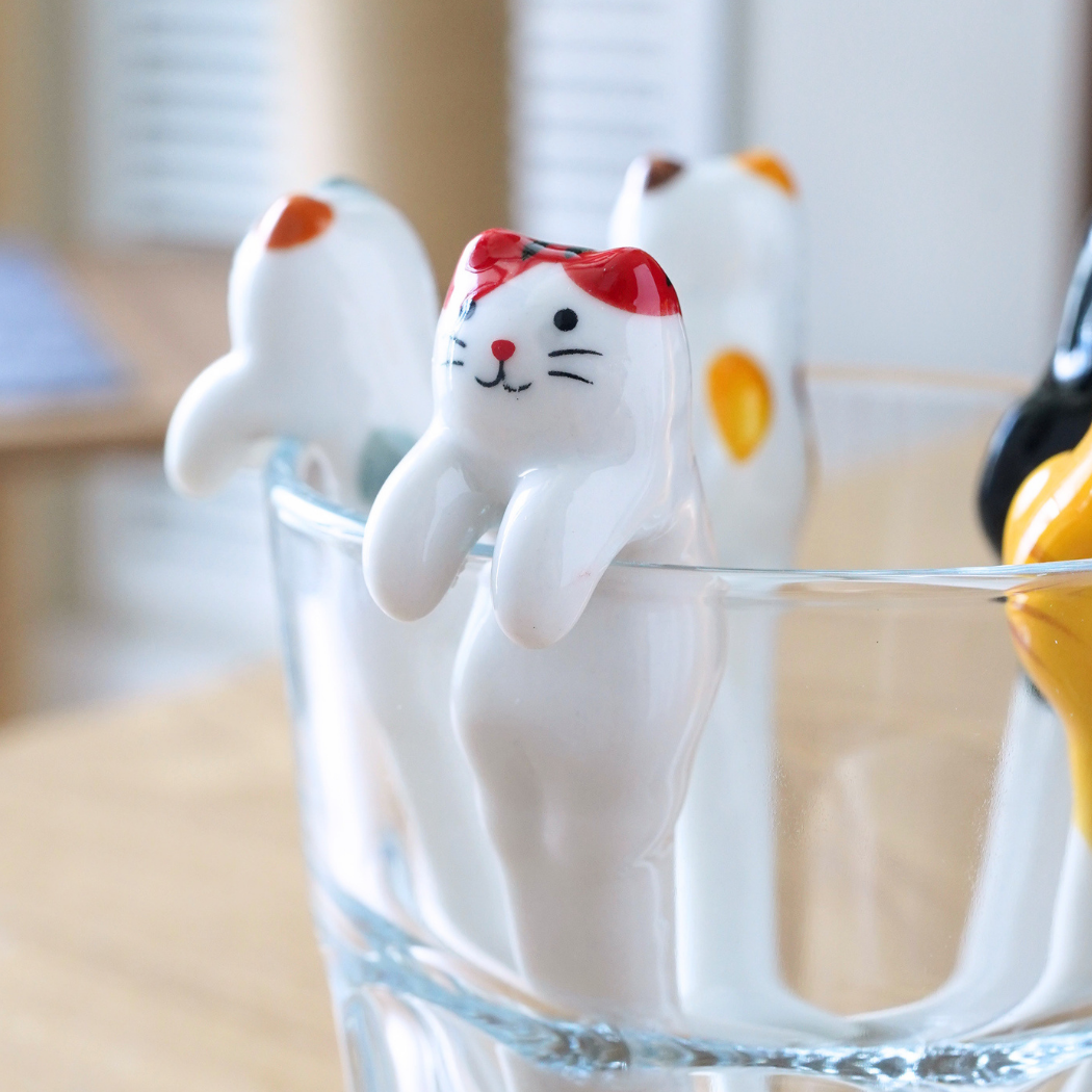 A set of cat-shaped plastic utensils, including a white cat with a red bow, a white cat with an orange spot, a black cat, and a yellow cat, arranged around the rim of a clear glass on a wooden table.