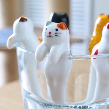 Three cat-shaped utensils or tools with painted faces are arranged inside a clear glass on a wooden surface, with a softly blurred indoor background.