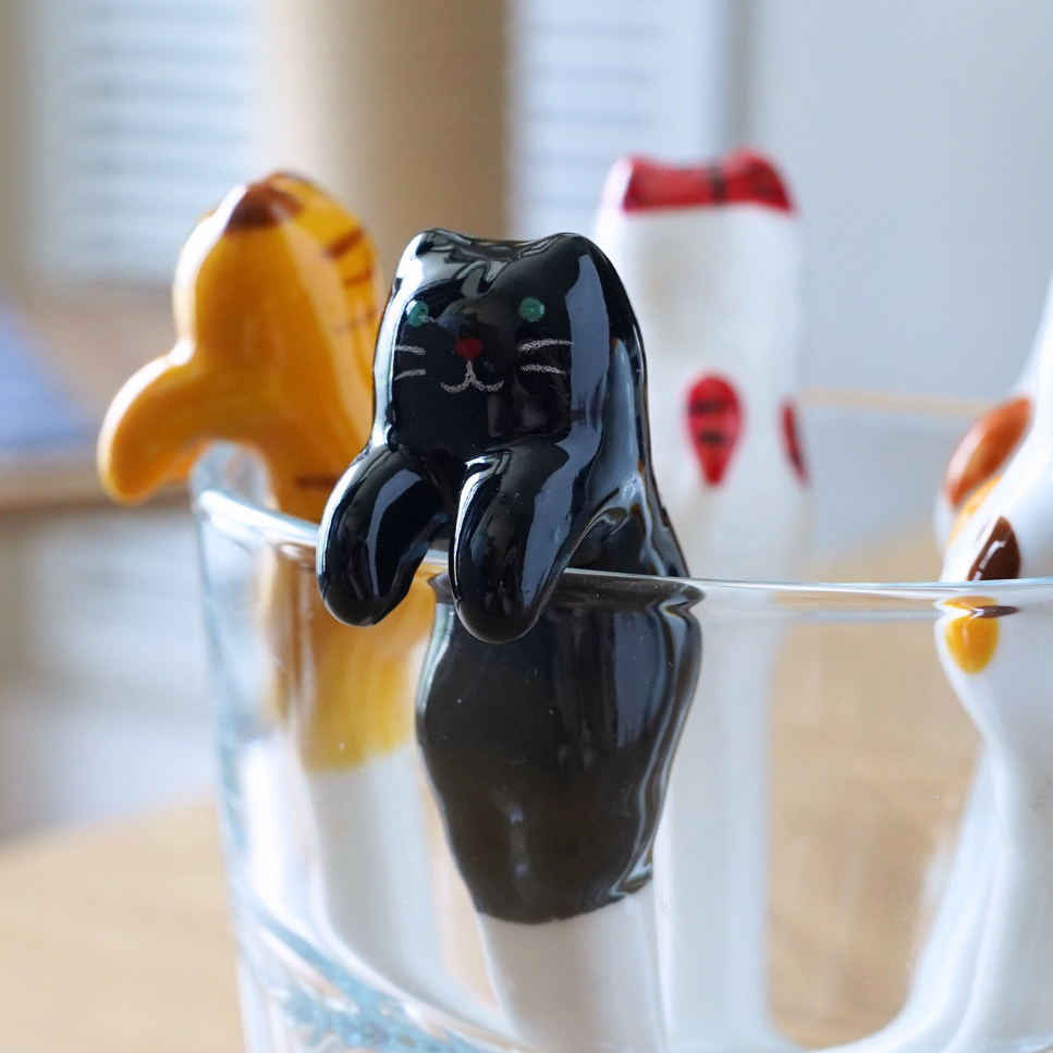 Three small cat-shaped utensils or tools in glossy black, white, and orange-yellow are arranged around the rim of a clear glass on a wooden table, with a softly blurred indoor background.