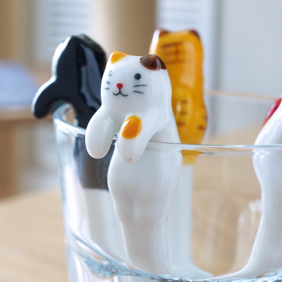 Three small cat-shaped utensils or tools resting inside a clear glass on a wooden table, with a softly blurred indoor background.