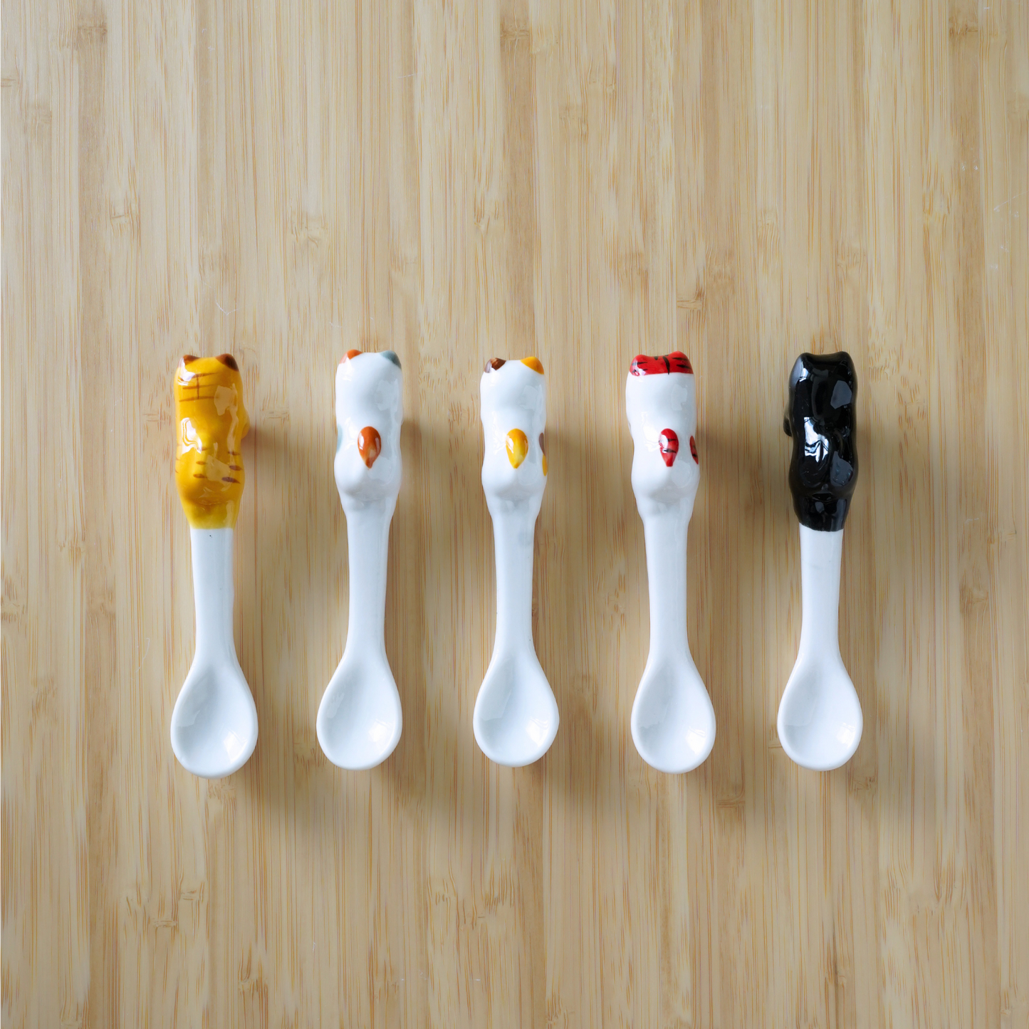 Five ceramic condiment spoons arranged in a row on a light wood background. Each spoon has a white bowl and handle, with the top of the handle decorated to resemble a dollop of sauce in different colors: mustard yellow, orange, yellow, red, and black.