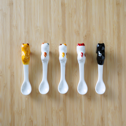 Five ceramic condiment spoons arranged in a row on a light wood background. Each spoon has a white bowl and handle, with the top of the handle decorated to resemble a dollop of sauce in different colors: mustard yellow, orange, yellow, red, and black.