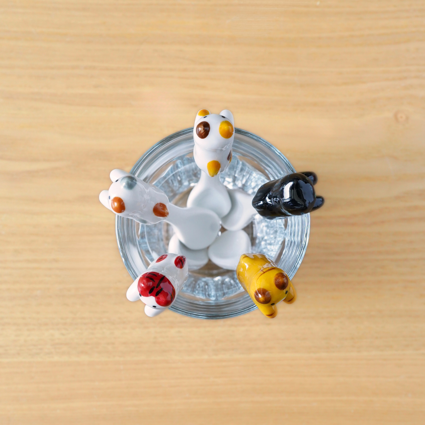 Four small ceramic animal figurines in white, black, yellow, and red are arranged around a clear glass cup on a light wood surface.