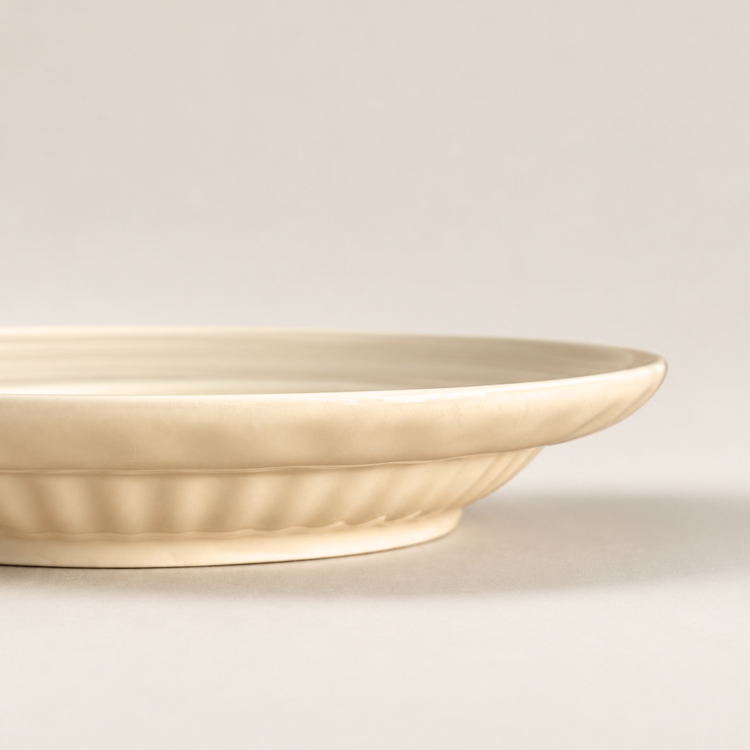 A close-up side view of a cream-colored ceramic plate with a fluted rim, set against a neutral light gray background.