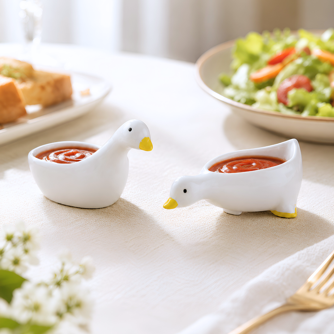 Two white duck-shaped sauce containers filled with red ketchup on a light beige tablecloth, with a salad bowl and bread plate blurred in the background.
