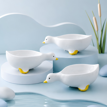Three white duck-shaped ceramic bowls with yellow beaks and feet, displayed on light blue pedestals with a soft blue studio background and decorative reeds.