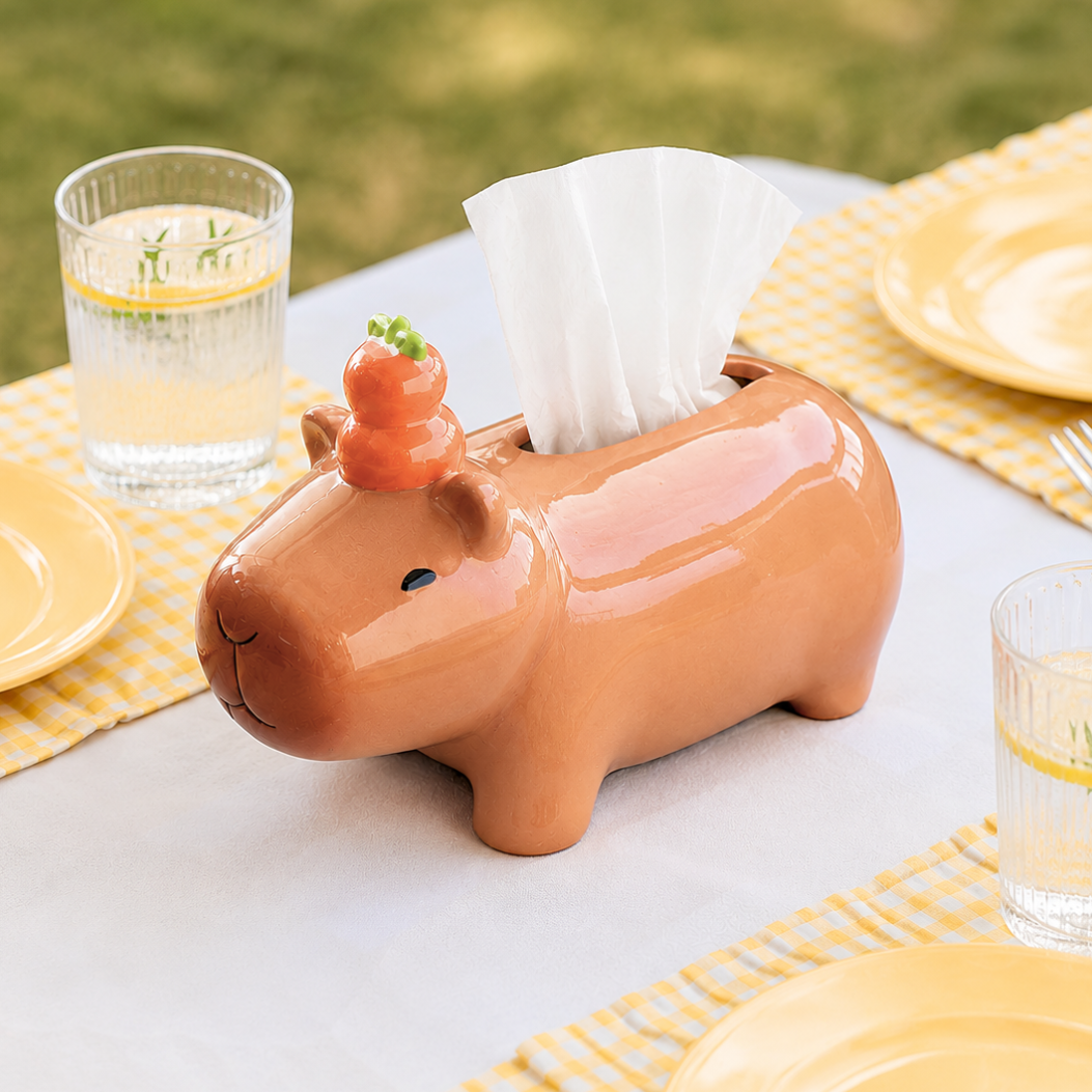 A glossy ceramic tissue box cover shaped like a capybara, with an orange yuzu on its head and white tissue paper partially pulled out, set on an outdoor table with yellow plates and glasses.