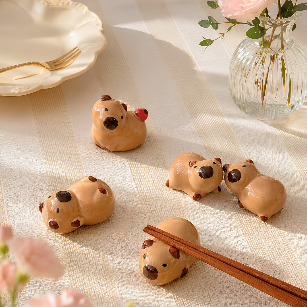 Five small ceramic animal capybara figurines or chopstick rests in a warm beige table cloth, arranged on a dining table with rose and flowers in case. White plates on the dining with a fork.