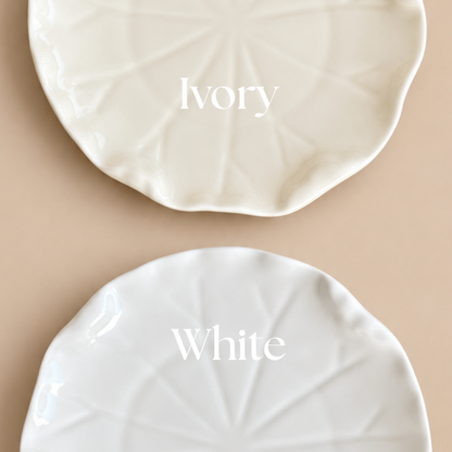 Two round ceramic plates with a leaf-like radial pattern, shown on a beige background. The top plate is labeled 'Ivory' and appears warm off-white, while the bottom plate is labeled 'White' and appears bright white.
