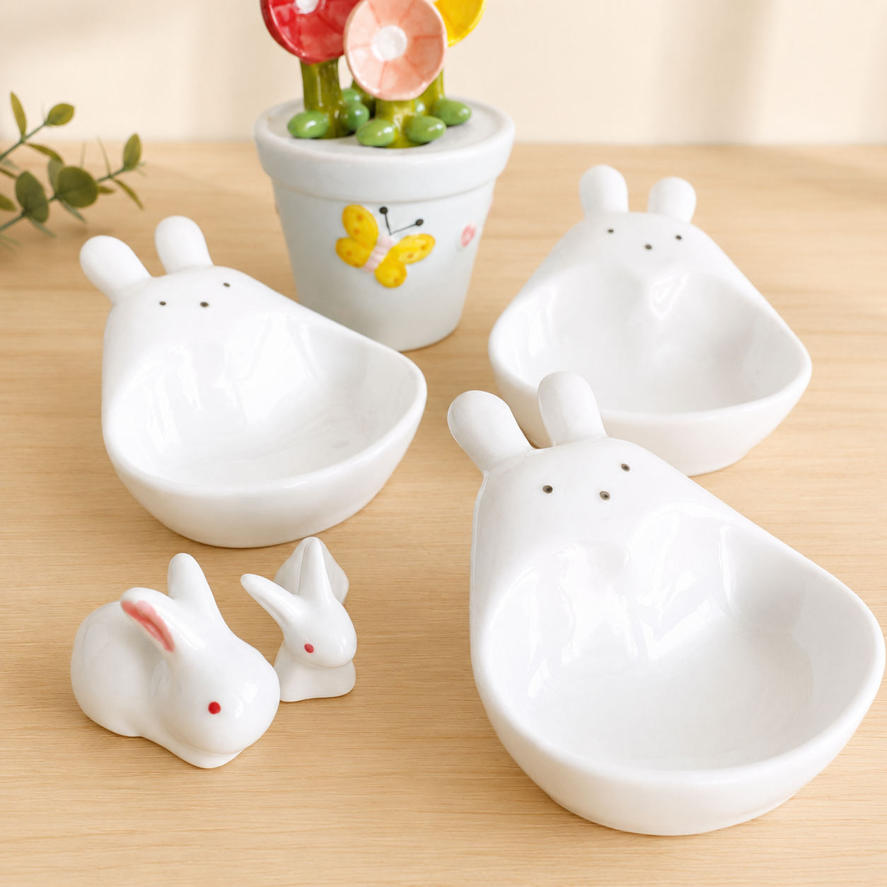 A set of white ceramic rabbit-shaped bowls and small rabbit figurines on a light wood surface, with a small potted flower in the background.