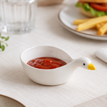 A white ceramic duck-shaped dipping bowl filled with ketchup, set on a beige tablecloth with a blurred dining background showing a burger, fries, and a glass.