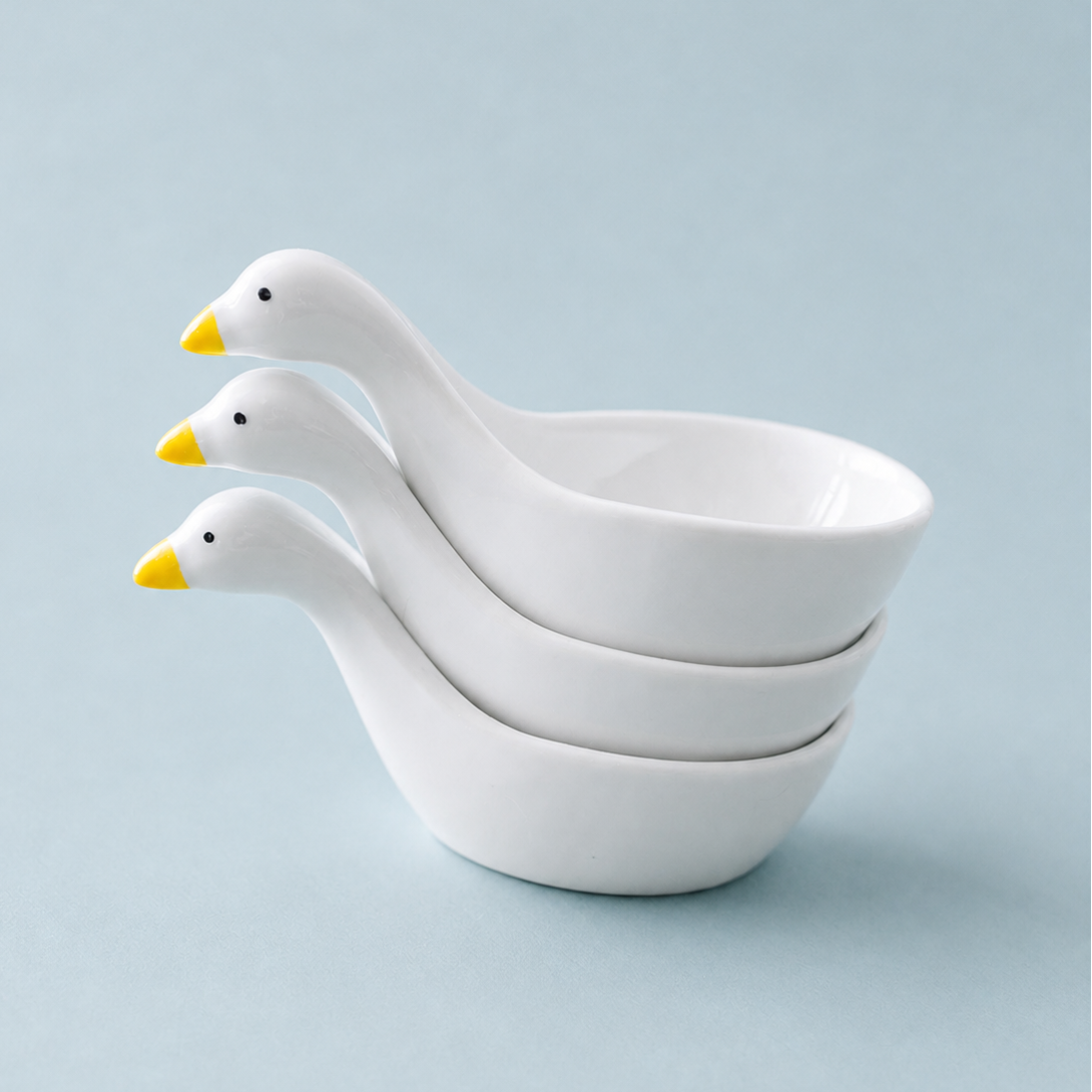 Three stacked white ceramic duck-shaped bowls with yellow beaks on a light blue background.