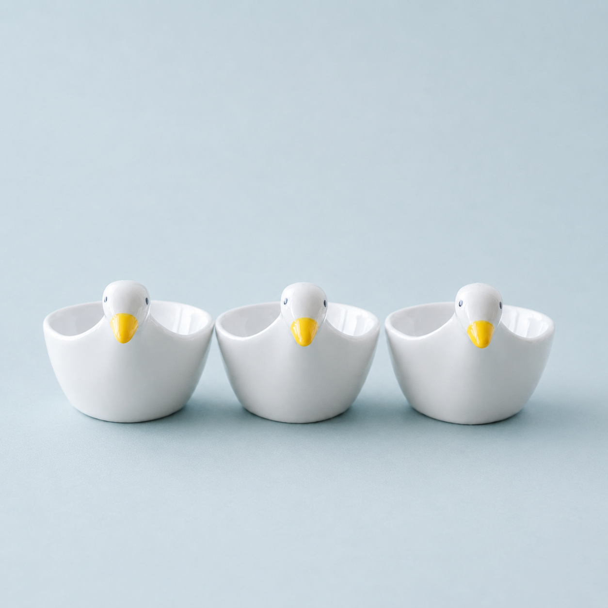 Three small white ceramic duck-shaped bowls with yellow beaks, arranged side by side on a light blue background.