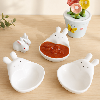Three white ceramic bunny-shaped bowls with small black eyes and noses are arranged on a light wood surface. One bowl contains red sauce, and a small decorative flower pot sits in the background.