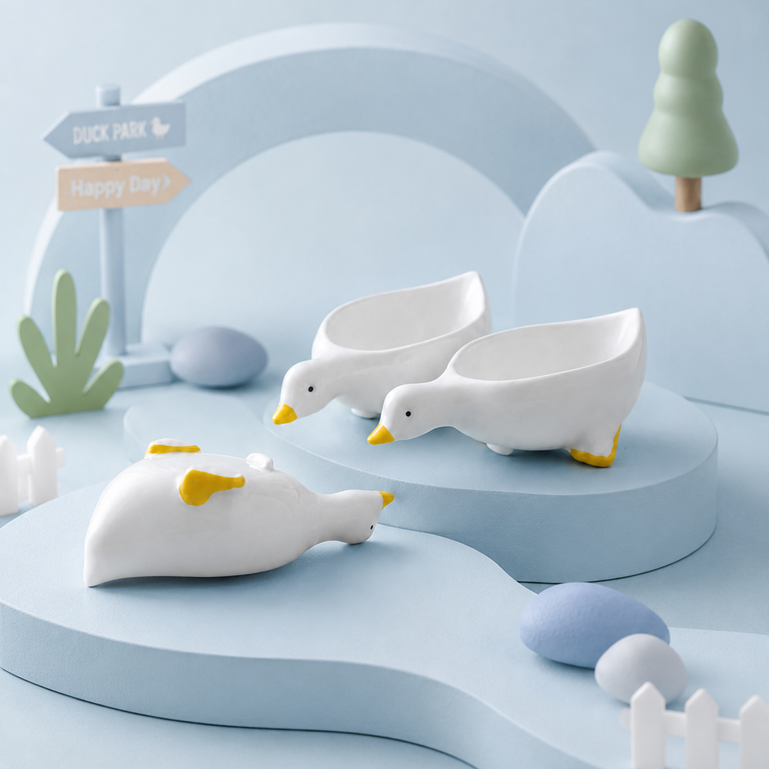 Three white duck-shaped bowls with yellow beaks and yellow webbed feet, styled on a pastel blue platform with decorative landscape props including a small tree, signpost, and stones.