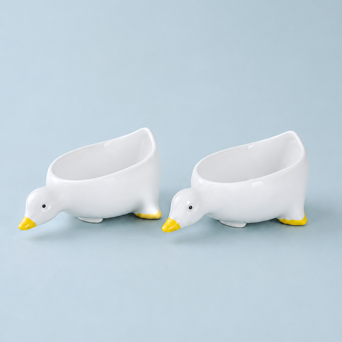 Two white ceramic duck-shaped bowls with yellow beaks and feet, arranged side by side on a light blue background.