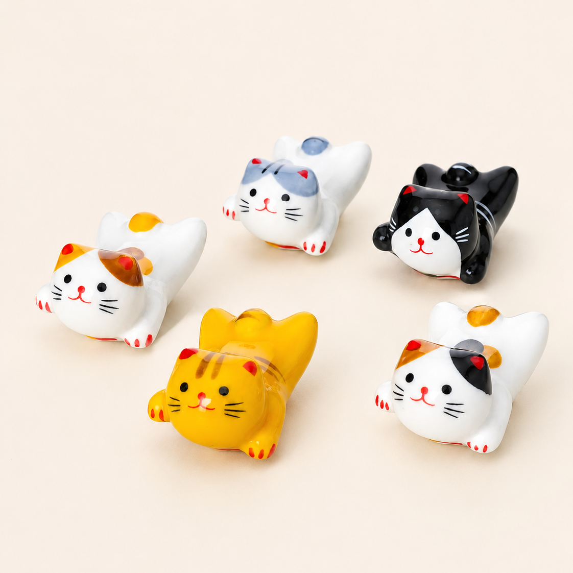 Five small cat figurines in different colorways arranged on a plain warm beige background. The cats include white, black, yellow, and calico designs with simple facial features.