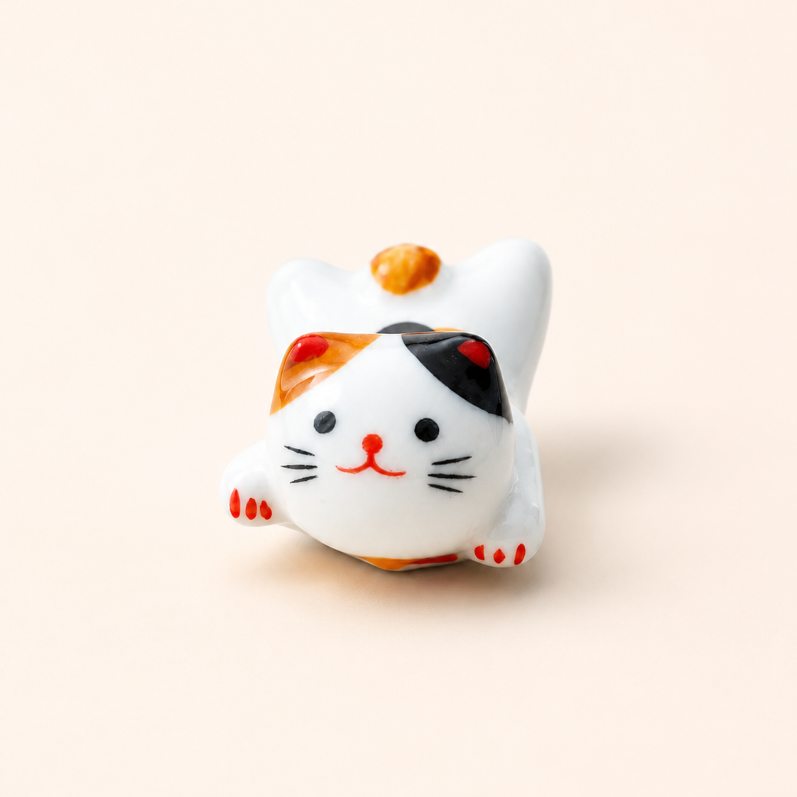 A small ceramic maneki-neko figurine with a white body, black and orange patches, and red details, posed on a light beige background.