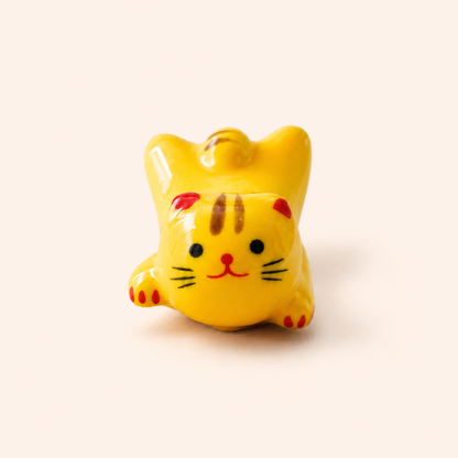 A small glossy yellow cat charm or pendant with red, black, and brown painted details, photographed on a plain warm off-white background.