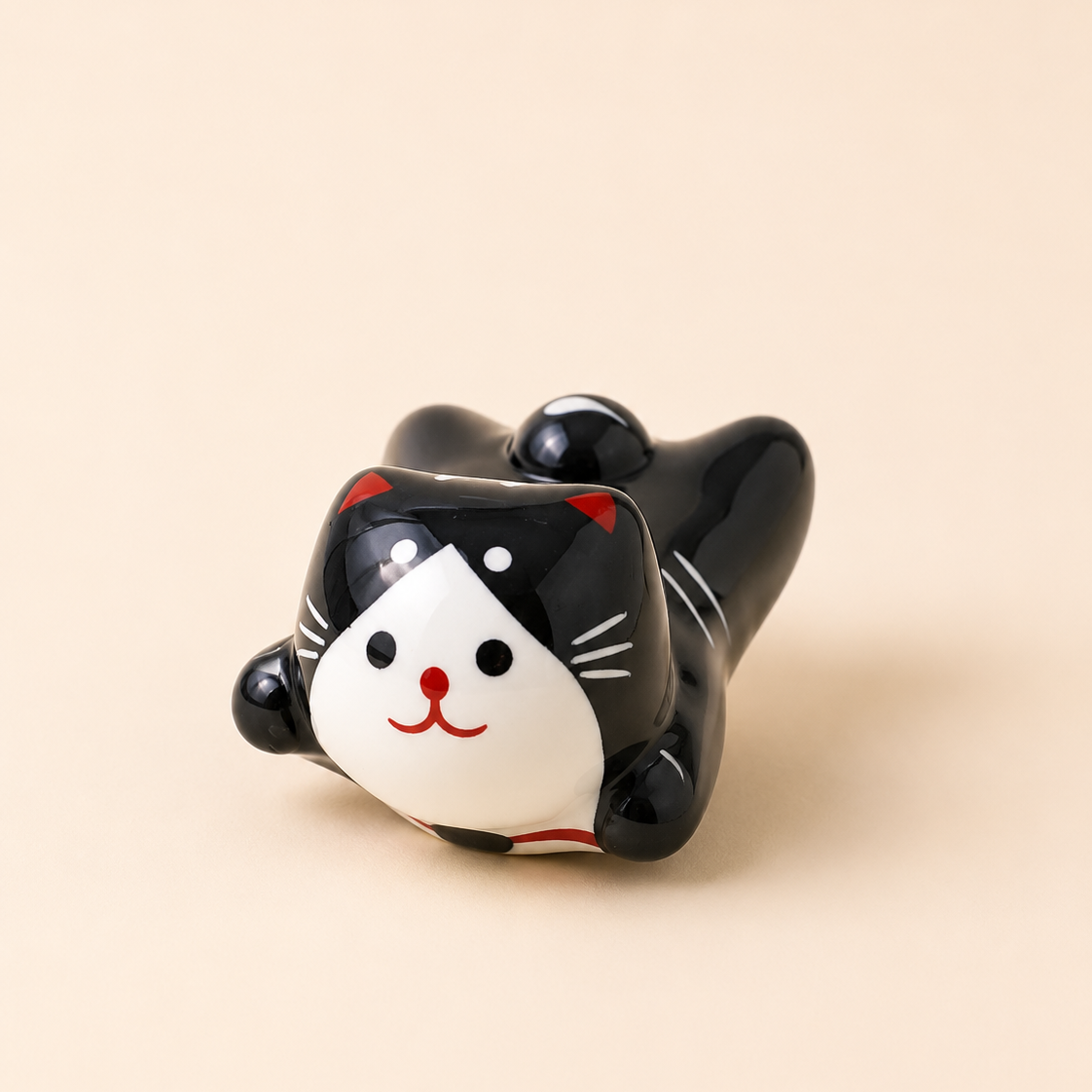 A glossy ceramic cat figurine lying down, featuring a black body, white face, red nose and whiskers, and small red ear accents, set against a solid beige background.