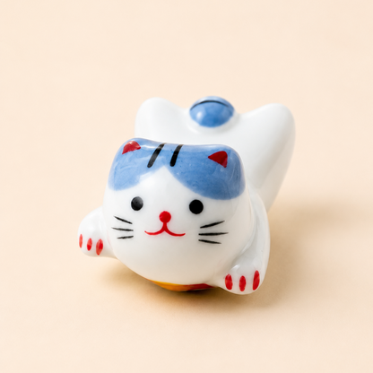 A small white ceramic lucky cat figurine with blue, red, and black painted details, photographed on a plain warm beige background.