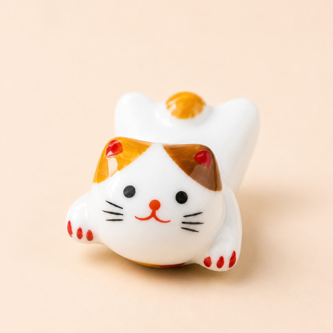 A small ceramic lucky cat figurine with a white body, orange patches, and red accents, posed on a warm beige background.