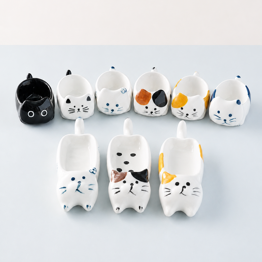 A set of ceramic cat-shaped bowls arranged on a light blue-gray surface. The collection includes solid black, white, and multicolor hand-painted cat bowls in various designs.