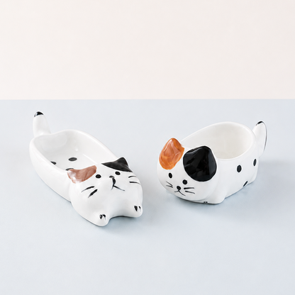 Two ceramic cat-shaped bowls shown on a light blue surface against a white background. One bowl is lying down in white with black spots and a black nose, and the other is sitting up in white with a black face, black spots, and an orange ear.