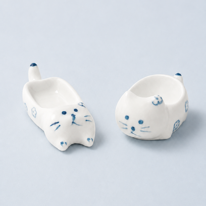 Two small white ceramic cat-shaped dishes with blue hand-painted details, shown on a light blue-gray background.