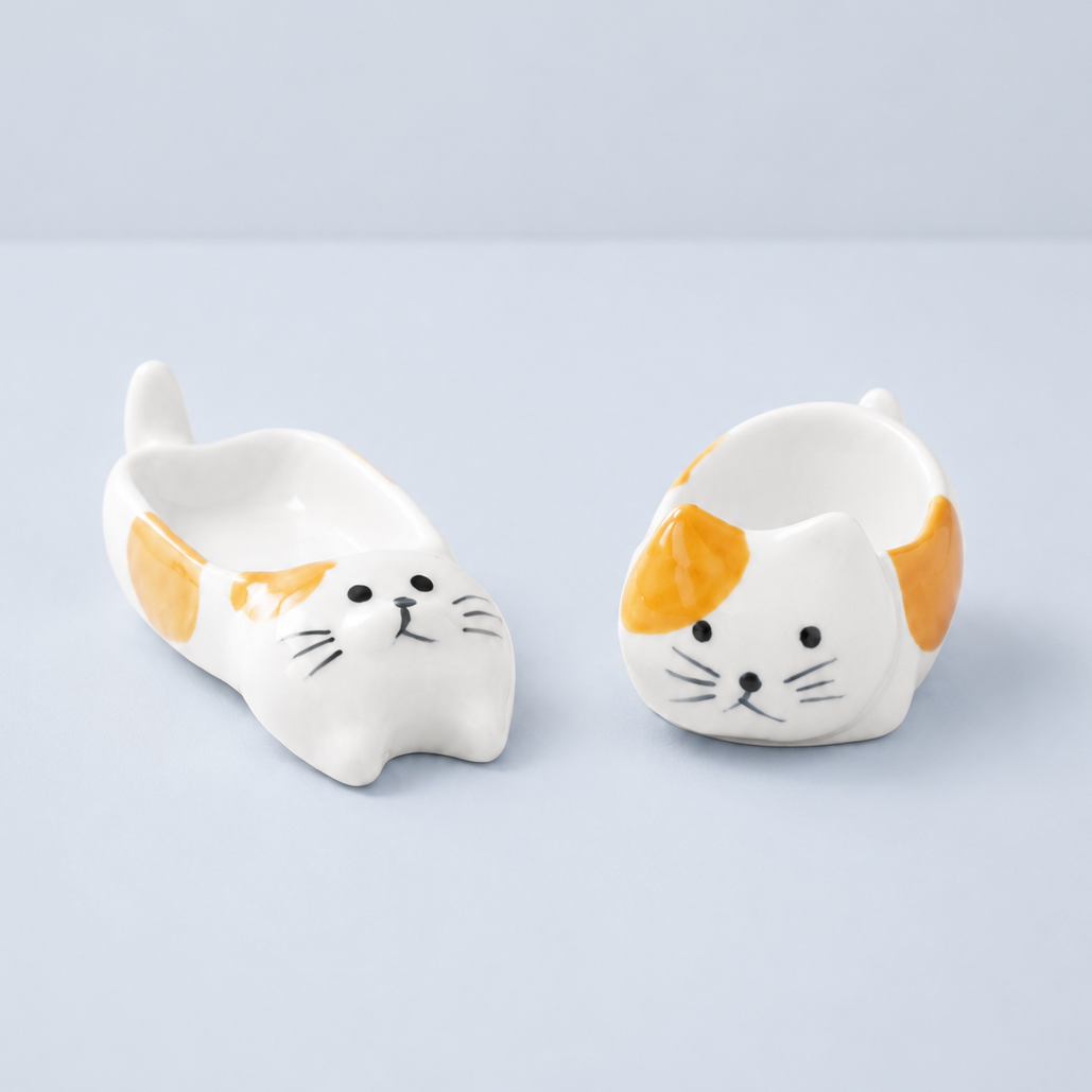 Two small white ceramic cat-shaped dishes with orange patches and black whisker details, shown on a light gray studio background.