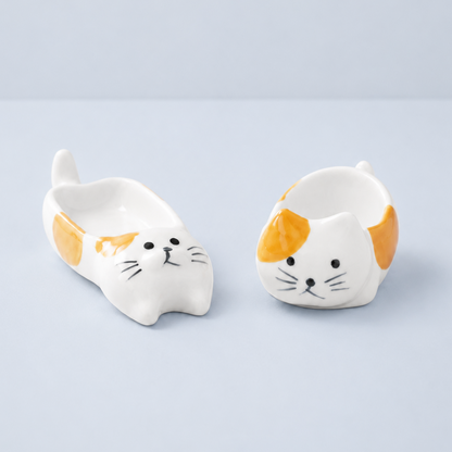 Two small white ceramic cat-shaped dishes with orange patches and black whisker details, shown on a light gray studio background.
