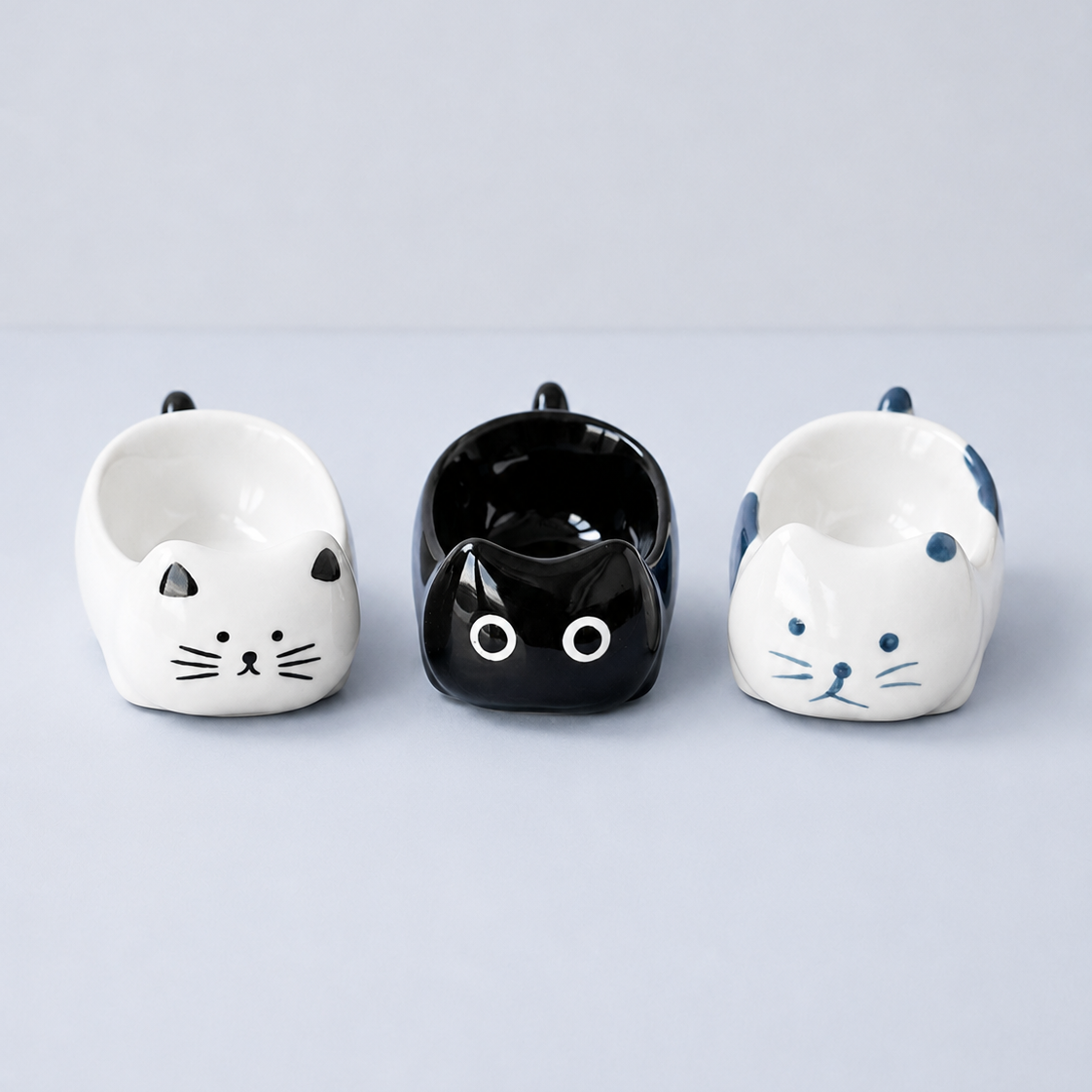 Three ceramic cat-shaped bowls in white, black, and white with blue accents, arranged side by side on a light gray background.