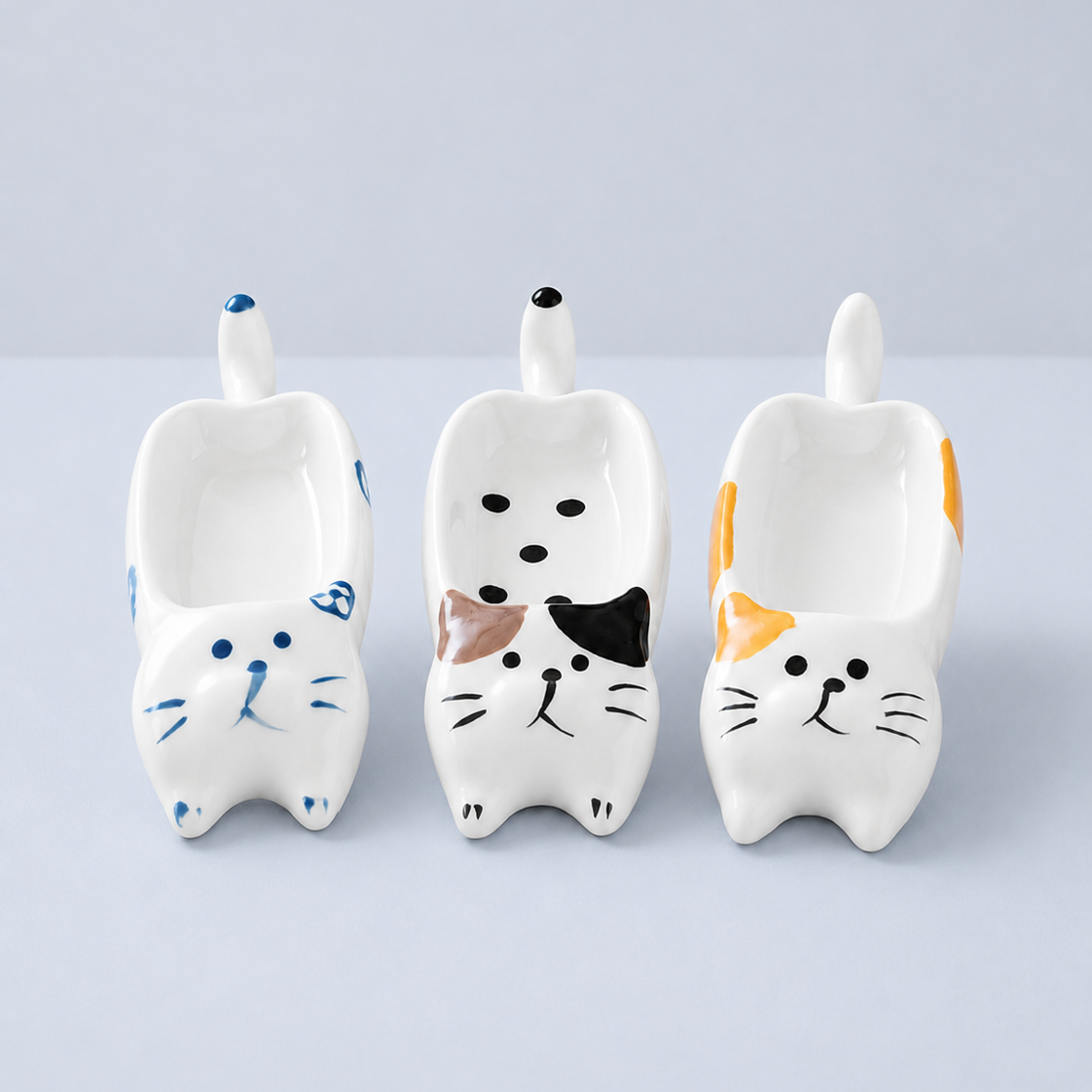 Three ceramic cat-shaped bowls in white, decorated with blue, black, brown, orange, and yellow painted details, arranged side by side on a light gray background.