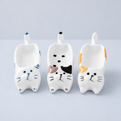 Three ceramic cat-shaped bowls in white, decorated with blue, black, brown, orange, and yellow painted details, arranged side by side on a light gray background.