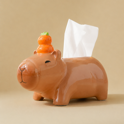 A glossy terracotta-colored capybara-shaped tissue box holder with a small orange yuzu on its head and a white tissue paper emerging from the back, set against a solid beige background.