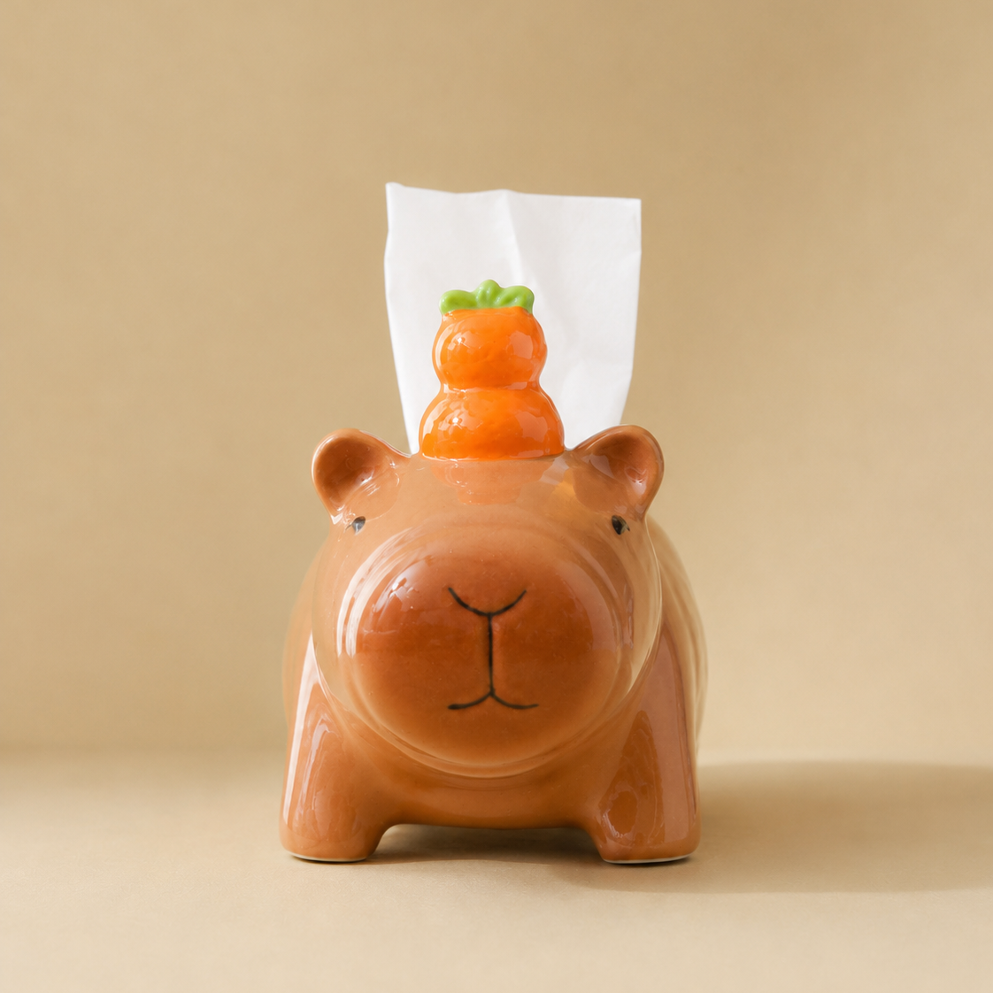 A ceramic tissue box cover shaped like a capybara in a warm terracotta brown, with a small orange yuzu decoration on top and a white tissue protruding. The product is photographed against a solid, warm beige background.