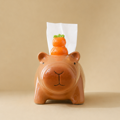 A ceramic tissue box cover shaped like a capybara in a warm terracotta brown, with a small orange yuzu decoration on top and a white tissue protruding. The product is photographed against a solid, warm beige background.