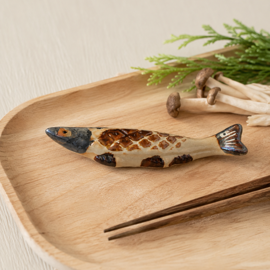 A small fish-shaped ceramic chopstick rest or utensil with a mottled cream, brown, and dark blue glaze, resting on a light wood tray with chopsticks and mushrooms nearby.