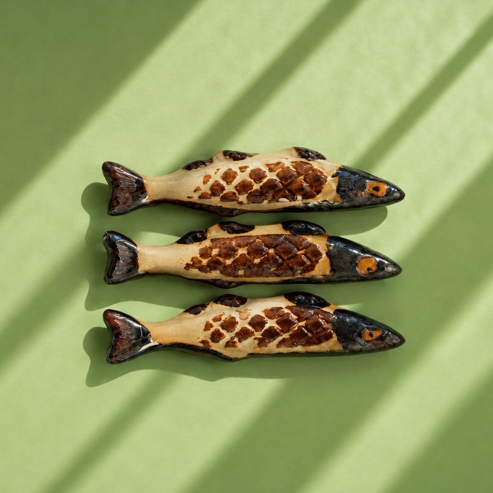 Three decorative fish-shaped chopstick rests or ornaments arranged vertically on a light green background with diagonal shadow stripes. The fish have cream bodies with dark brown and black spotted scales, dark heads, and orange eyes.