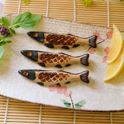 Three decorative fish-shaped ceramic chopstick rests arranged on a rectangular ceramic plate with pink cherry blossom motifs, garnished with green leaves and lemon slices, set on a bamboo mat.