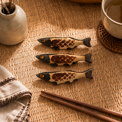 Three small fish-shaped ceramic chopstick rests with a brown and cream patterned scale design are arranged on a woven straw placemat next to wooden chopsticks, a small vase with dried flowers, and a cup of tea.