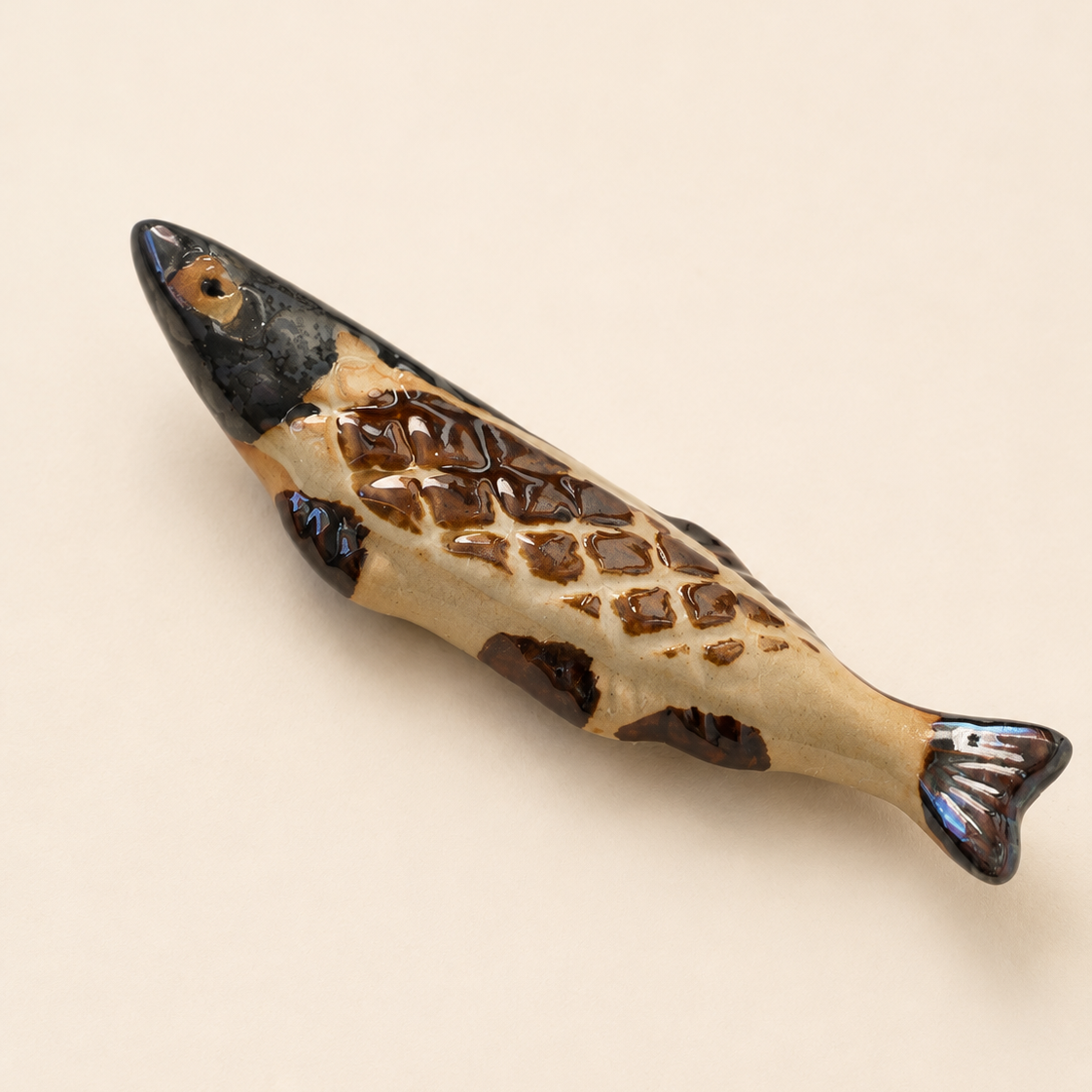 A ceramic fish figurine or chopstick rest with a glossy finish, featuring a beige body with brown scales and a dark black head, resting on a solid light beige background.