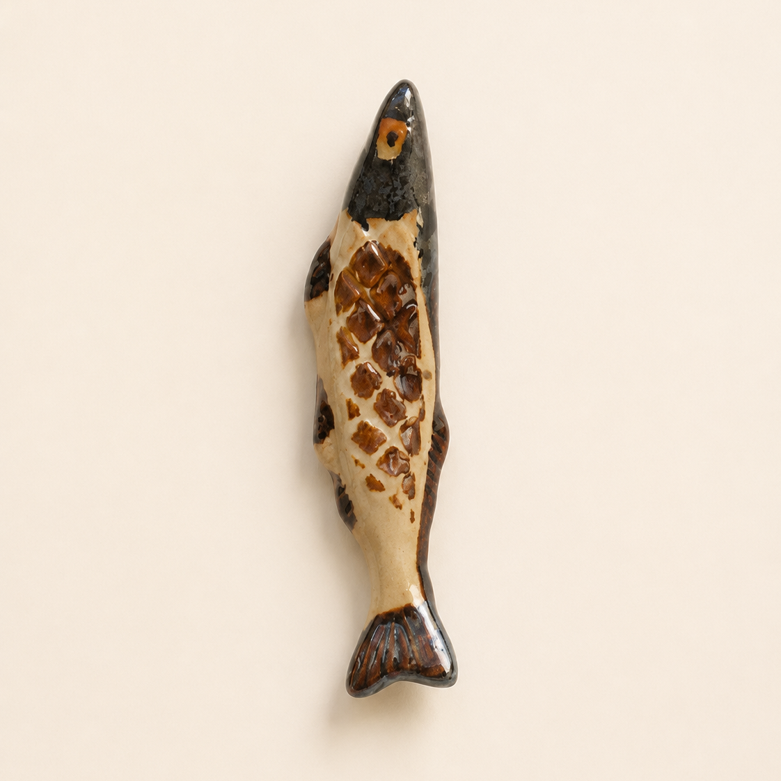 A small fish-shaped decorative object and chopstick rests with a glossy finish, featuring a cream-colored body with brown scale patterns and a dark gray to black head and fins, set against a solid light beige background.
